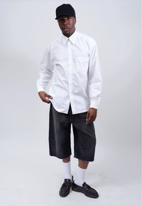 White button-up shirt with long sleeves, black denim shorts, white crew socks, and black shoes. Model wears a black cap, neutral background.