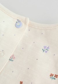Vêtement pêche pâle avec de petits imprimés floraux en violet, rose et vert ; comporte une fermeture à boutons au niveau de l'encolure avec un motif de fleur pastel.