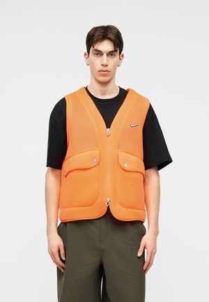 Mladý muž v oranžovej zipsovej veste s dvoma chlopňovými vreckami, oblečený na čiernom tričku a olivových zelených nohaviciach, stojí pred jednoduchým pozadím.