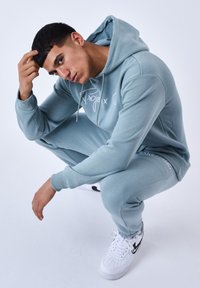 Haut à capuche bleu clair et pantalon de jogging assorti en tissu doux ; comporte un logo sur la poitrine. Porté avec des baskets blanches.