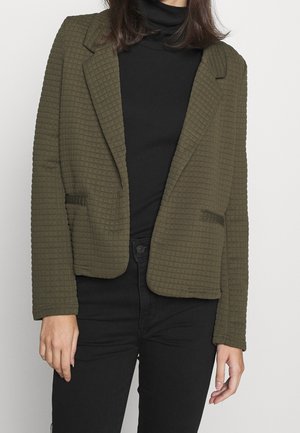 Kvinde iført en tekstureret olivengrøn blazer over en sort højhalset trøje og sorte bukser, stående mod en ensartet lys baggrund.
