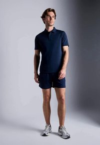Jovem de pé, vestindo uma camisa polo azul-marinho, calções combinando e sapatos de desporto brancos, contra um fundo cinza liso.