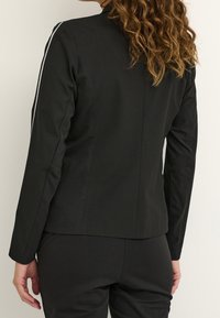 Veste noire à manches longues, coupe ajustée, avec des bandes blanches sur les côtés. Confectionnée dans un tissu lisse avec un design épuré et minimaliste.