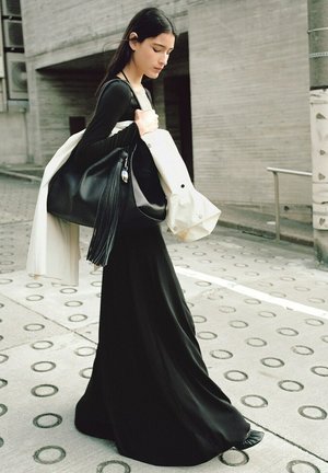 Młoda kobieta w długiej czarnej sukience i aparacie słuchowym, niosąca białą kurtkę i dużą czarną torbę, spacerująca po wzorzystym chodniku.