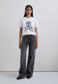 Donna in piedi che indossa una maglietta bianca con grafica di teschio e ossa incrociate, pantaloni a gamba larga neri sbiaditi e scarpe nere su uno sfondo semplice.