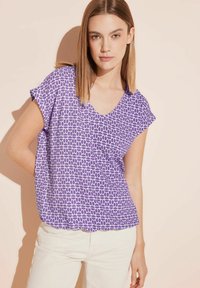 Top corto viola a maniche corte con scollo a V e motivo geometrico in viola scuro. Tessuto morbido con vestibilità rilassata, abbinato a pantaloni bianchi.