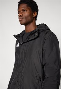 Veste noire à capuche avec une texture lisse et brillante, dotée d'une fermeture éclair et d'un logo Adidas blanc sur le côté gauche de la poitrine.