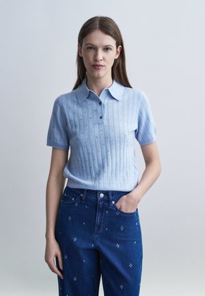 Femme aux longs cheveux bruns portant un polo en maille à manches courtes bleu clair et un jean bleu foncé avec de petits motifs brodés blancs.