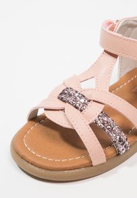 Różowe sandały dla malucha z błyszczących paskiem, otwartym czubkiem, wyściełaną brązową podeszwą i rzepem przy kostce na białym tle.