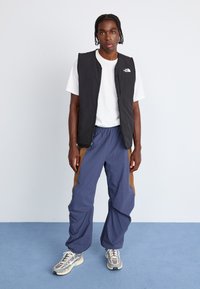 Sort ærmeløs vest, hvid t-shirt under, blå bukser med brune detaljer, og grå sneakers med netpaneler og reflekterende elementer.