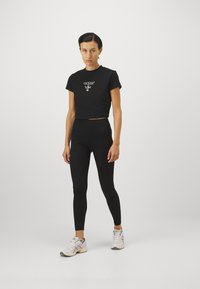 Crna kratka majica s bijelim logotipom "GUESS", uparena s crnim tajicama. Model nosi bijele sportske tenisice. Jednostavan, prilagođen dizajn.