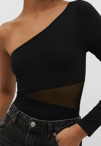 Bodysuit noir avec un design à une épaule, comportant un panneau en maille et une longue manche. La texture est lisse et moulante, associée à un jean foncé.