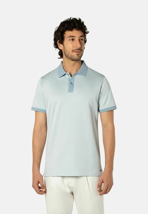 Man met krullend donker haar, gekleed in een lichtblauw poloshirt met een donkerblauwe kraag en witte broek, staand tegen een neutrale achtergrond.