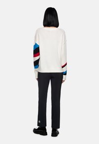 Weißer Pullover mit blauen, schwarzen und roten Streifenakzenten; gerippte Textur; kombiniert mit schwarzen Hosen; Modell von hinten betrachtet.