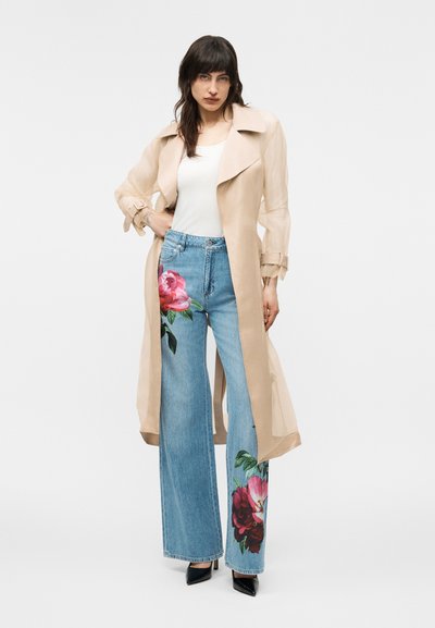 Femme portant un long trench-coat beige, un top blanc, un jean bleu à jambes larges avec de grands imprimés floraux et des talons noirs pointus sur fond uni.