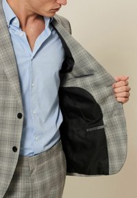 Chaqueta de traje a cuadros grises con forro negro, presentada abierta. Camisa de color azul claro debajo, con botones y cuello. Notable bolsillo interior.