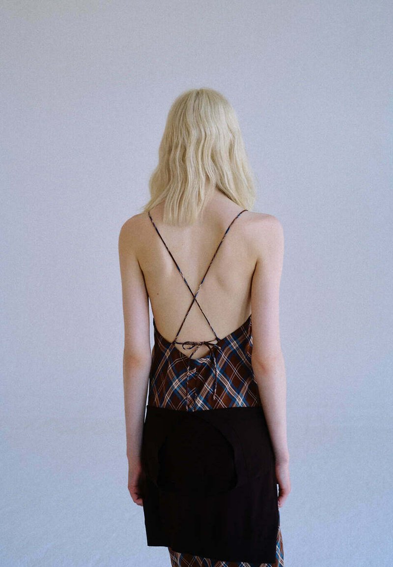 Femme blonde aux cheveux ondulés portant une robe à carreaux dos nu avec de fines bretelles croisées et un vêtement noir noué autour de sa taille.