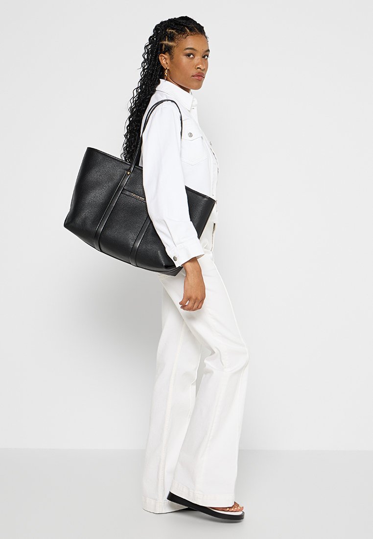 Schwarze Leder-Umhängetasche mit strukturiertem Finish, ausgestattet mit zwei langen Griffen und einer strukturierten Form, getragen von einer Person in weißer Kleidung.