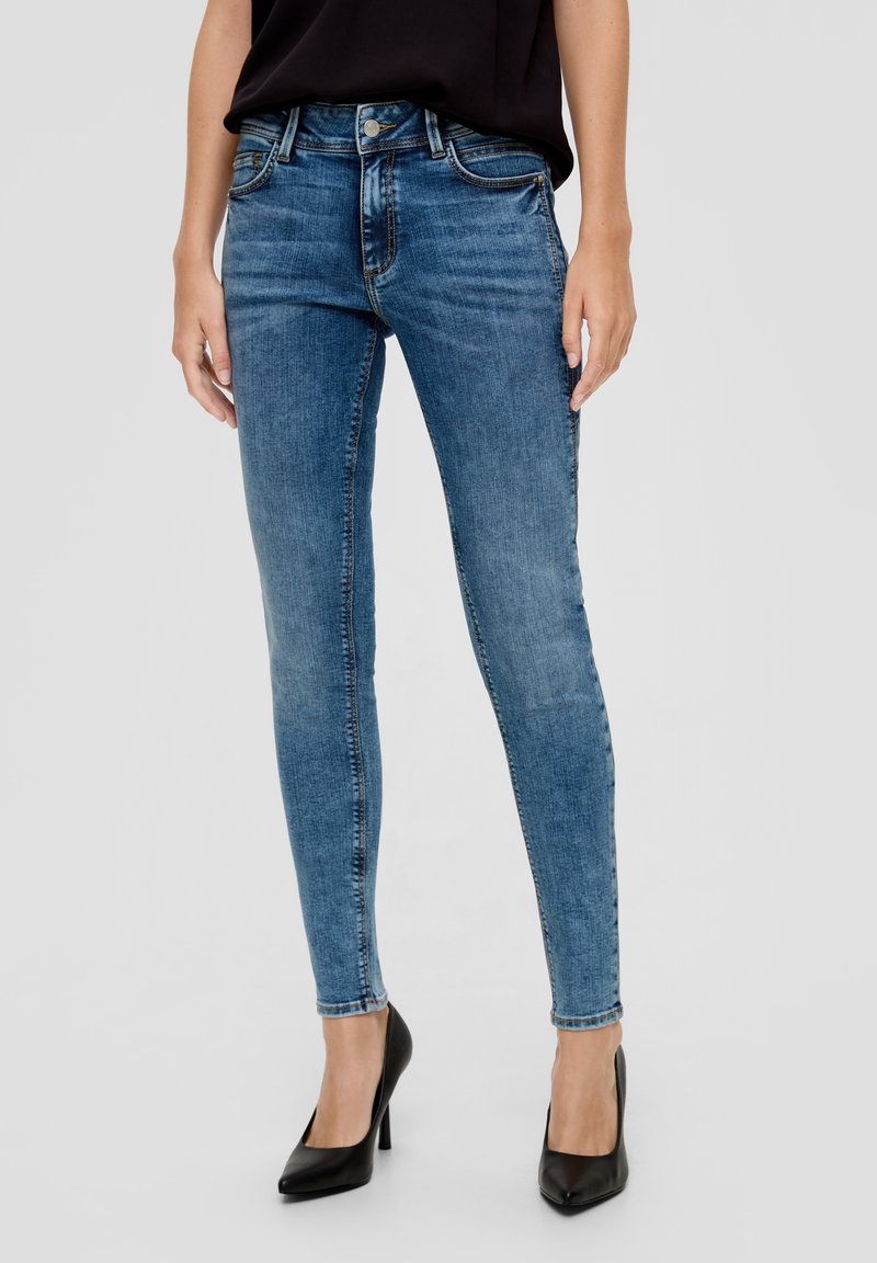 Jean skinny en denim bleu moyen avec un lavage délavé, conception à cinq poches, taille mi-haute et surpiqûres contrastantes ; associé à des talons hauts noirs.