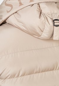 Giacca imbottita beige con finitura lucida, dotata di cappuccio, fibbia della cintura e grande logo in rilievo sulla parte superiore.