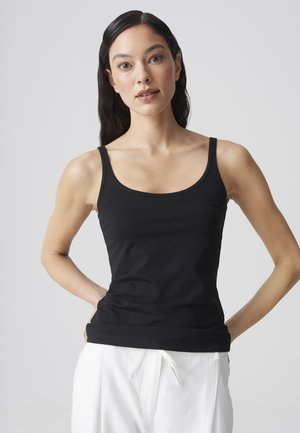 Vrouw met lang donker haar, gekleed in een zwarte mouwloze top en witte broek, staand met handen in de zij tegen een effen achtergrond.