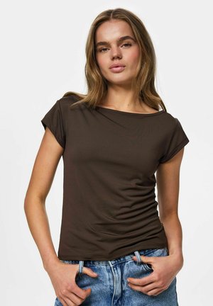 Giovane donna con capelli castano chiaro che indossa una maglietta a maniche corte marrone scuro e jeans di denim blu, in posa con le mani nelle tasche.