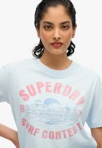 Camiseta de manga corta azul claro con una impresión gráfica en rojo y azul, que representa una escena de surf con el texto "SUPERDRY SURF CONTEST."