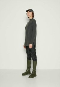 Graues langärmliges Stricktop, schwarze Slim-Fit-Hosen und grüne knielange Gummistiefel. Akzentuiert mit einer dunklen Mütze und minimalistischem Schmuck.