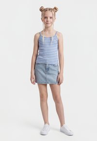 Niña joven con dobles moños trenzados, vestida con una camiseta de tirantes azul a rayas, falda de mezclilla y zapatillas blancas, de pie sobre un fondo blanco.