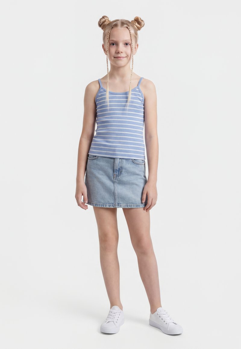 Niña joven con dobles moños trenzados, vestida con una camiseta de tirantes azul a rayas, falda de mezclilla y zapatillas blancas, de pie sobre un fondo blanco.