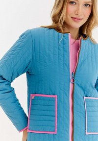 Niebieska pikowana kurtka z różowymi akcentami i kieszeniami. Posiada różowy zamek błyskawiczny i brązowy kołnierz. Teksturowany materiał z poziomym przeszyciem.