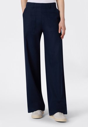 Pantalon large bleu marine à poches plaquées avant porté avec des baskets blanches sur un fond uni.