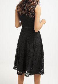 Svart spetsklänning med ärmlös design, har en figursydd bodice och en utsvängd kjol som faller under knäet. Texturerad blommönster genom hela.