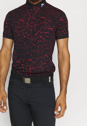 Mężczyzna w czarnej koszulce polo z czerwonym wzorem spękań, czarnym kołnierzem, czarnych spodniach i czarnym pasku z metalową klamrą.