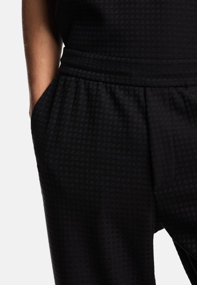 Jogging di colore nero con texture e pattern a scacchi, dotati di vita elastica e tasche laterali, mostrati dall'altezza della vita fino alla coscia.