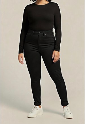 Personne portant un haut à manches longues noir, un jean taille haute noir avec les ourlets retroussés, et des baskets blanches, se tenant devant un fond uni.