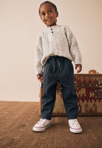 Grijze kabelgebreide trui met drie bruine knopen, marineblauwe geruite broek en witte sneakers met rode accenten, staand op een gestructureerde vloer.