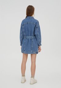 Veste en jean bleu clair à manches longues, taille ceinturée et poches. Dotée d'un col classique et d'une légère usure à l'ourlet.