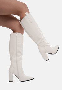 Botas blancas hasta la rodilla en material de brillo con estampado de cocodrilo, con tacón cuadrado, puntera puntiaguda y cremallera lateral para un fácil uso.