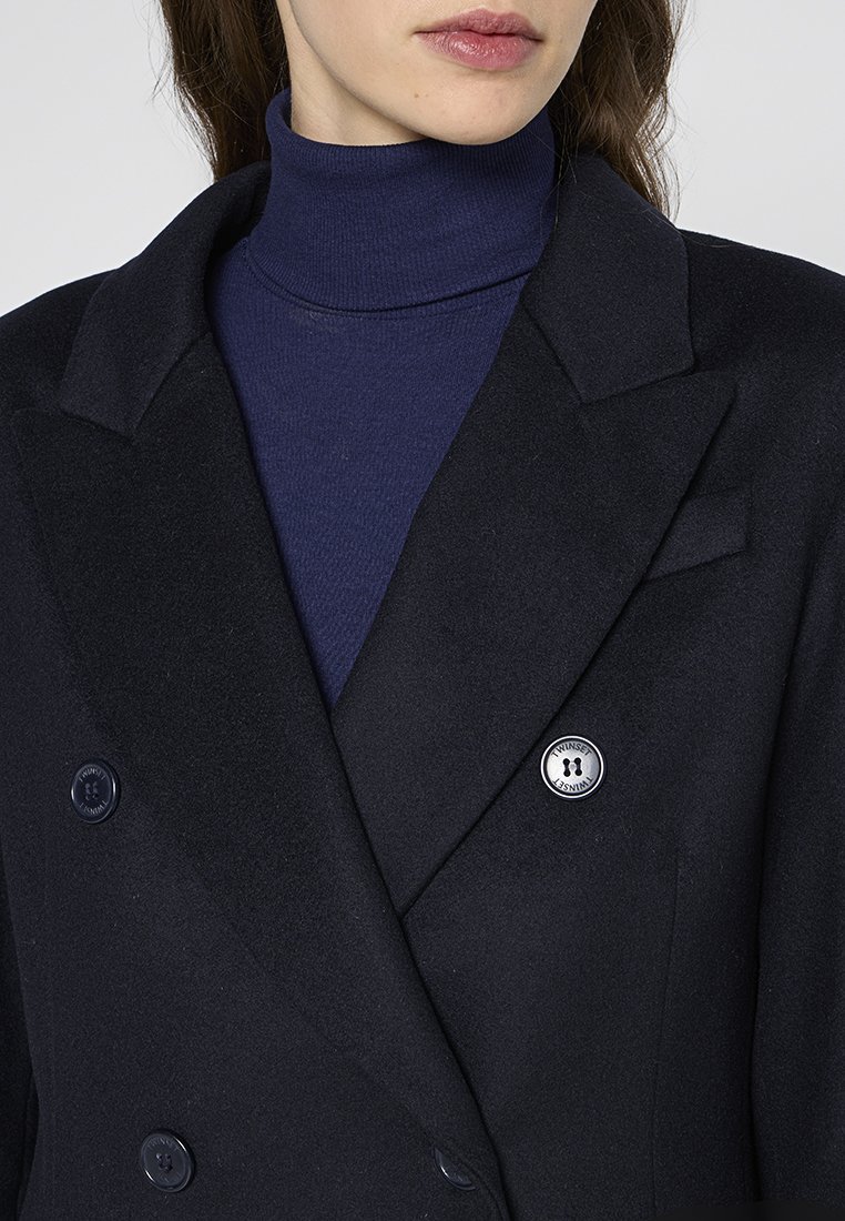 Dubbelzijdige zwarte wollen blazer van een wolmix met twee knopen, een reverskraag en een ronde logo-speld aan de linkerzijde. Marineblauwe coltrui eronder.