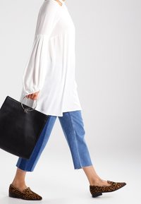 Vit långärmad topp, blå cropped jeans och bruna loafers med leopardmönster. En svart tote bag med ett runt handtag hålls i handen.