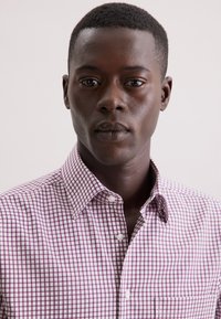 Bruine geblokte blouse met kleine paarse en witte vierkanten, klassieke kraag, knoopsluiting aan de voorkant en een enkele borstzak.