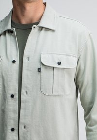 Lichtgrijze button-up shirt gemaakt van zachte stof, met twee borstzakken, zwarte knopen en een platte kraag. Gedeeltelijk over een groen shirt gedragen.