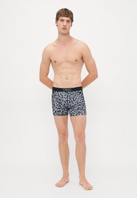 Ung mann står barføtt iført leopardmønstrede boxer shorts med svart livrem merket Versace på en enkel hvit bakgrunn.