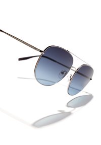 Ochelari de soare aviator cu ram�ă din metal și lentile cu un gradient albastru, aruncând umbre delicate pe o suprafață albă.