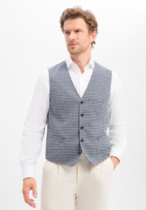 Mann trägt weißes Hemd und eine geknöpfte graue Hahnentrittweste mit beigen Hosen, steht mit einer Hand in der Tasche und schaut nach rechts.