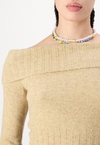 Beige strikkegenser med avkuttede skuldre og ribbestrikket hals og ermer, kombinert med en flerfarget perlekjede med smiley-fjes detaljer.