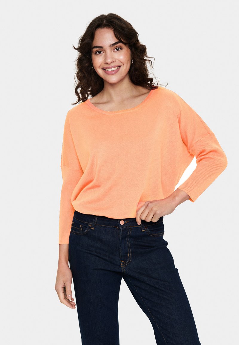 Leichter, lässig sitzender, korallfarbener Pullover mit weitem Halsausschnitt und 3/4-Ärmeln, kombiniert mit dunkelblauen Jeans. Glatte Textur.