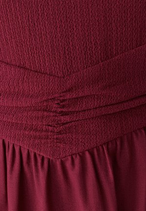 Robe bordeaux en tissu texturé, présentant des plis et des sections froncées pour le design. Tissu doux et fluide avec une finition lisse.