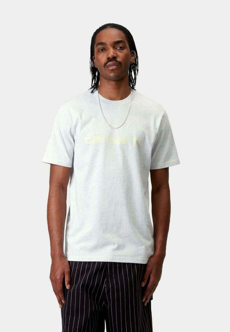 Camiseta de manga corta gris claro con el logo de "Carhartt" en un sutil amarillo. Se lleva con pantalones negros a rayas. Material de algodón, corte relajado.